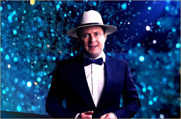 Arek Zasowski dressed in a tuxedo and fedora, standing against a blue cinematic bokeh background while hosting an episode of the Actors by Actors Show