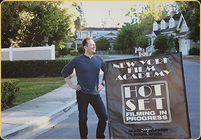 Arek Zasowski stands beside a New York Film Academy “Hot Set” sign during a filming workshop on the Universal Studios Hollywood backlot, wearing a navy long-sleeve shirt and jeans