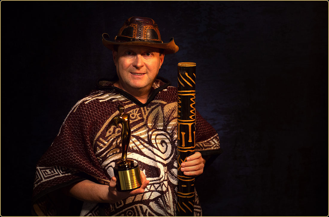 Award-winning filmmaker Arek Zasowski in a tribal poncho and adventure hat, proudly holding his Gold Telly Award and rainstick—evoking cinematic adventure and global storytelling